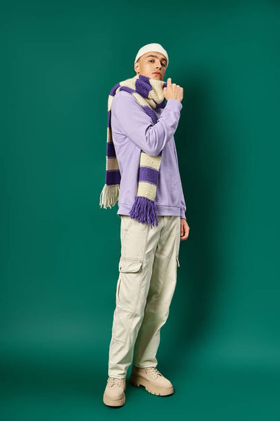 good looking man in purple sweatshirt, stripped scarf and beanie hat standing on turquoise backdrop - 写真・画像