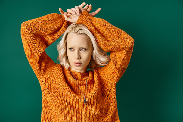 young blonde woman in mustard yellow sweater posing with hands above head on turquoise backdrop - Photo, Image