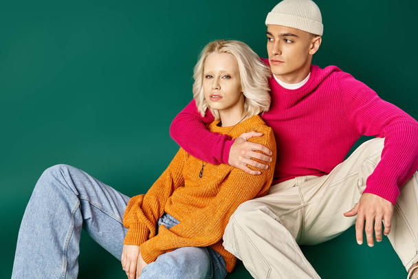 couple in winter sweaters, man in beanie hugging blonde woman while sitting together on turquoise - Photo, Image