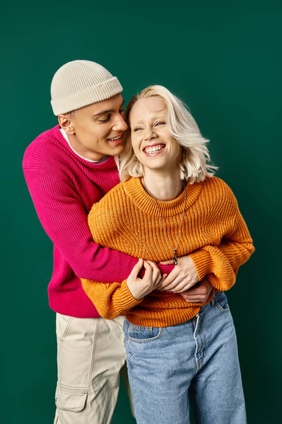 ζευγάρι σε χειμερινή ενδυμασία έχοντας τη διασκέδαση, ο άνθρωπος σε Beanie αγκαλιάζει ευτυχισμένη φίλη σε τυρκουάζ φόντο - Φωτογραφία, εικόνα