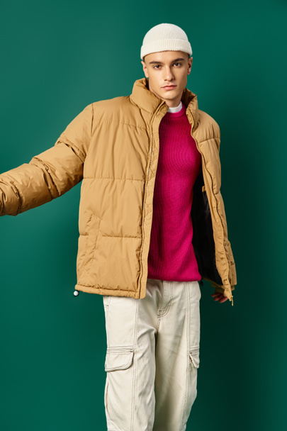 stijlvolle man in muts en winter kogelvrij vest poseren op turquoise achtergrond, winter trends - Foto, afbeelding