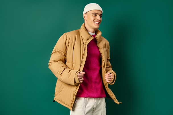 allegro giovane uomo in cappello berretto ed elegante giacca soffietto su sfondo turchese, tendenze invernali - Foto, immagini