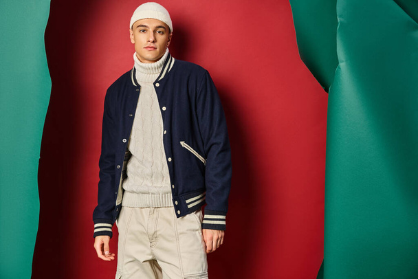 handsome man in beanie, white sweater and bomber jacket on ripped red with turquoise backdrop - Photo, Image