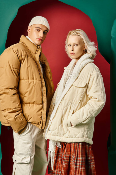 young couple in trendy winter jackets posing with hands in pockets on red with turquoise backdrop - Photo, Image