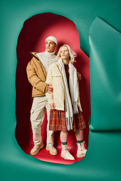 longitud completa de hombre y mujer con estilo en ropa de abrigo de invierno posando cerca de color turquesa desgarrado y fondo rojo - Foto, Imagen