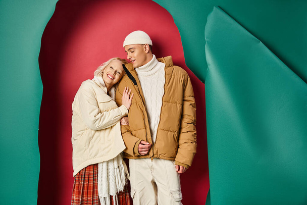 mujer complacida en chaqueta de invierno abrazando novio feliz cerca de desgarrado turquesa y fondo rojo - Foto, Imagen