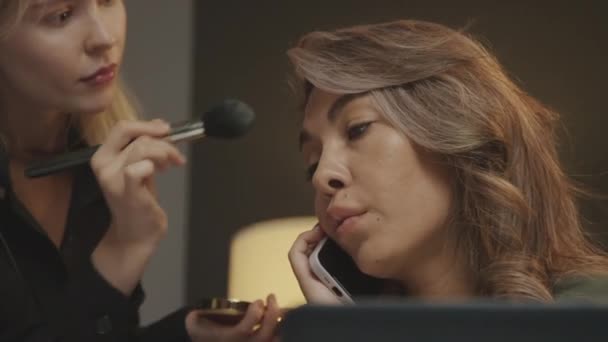Primer plano de artista de maquillaje caucásico aplicando polvo en la cara y las mejillas de la presentadora femenina, que está hablando en el teléfono inteligente, durante el descanso entre el rodaje en el estudio - Imágenes, Vídeo