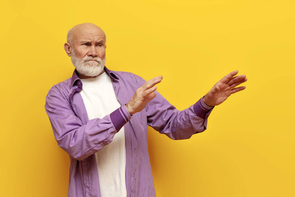 ontevreden oude kale grootvader met grijze baard vermijdt en negeert op gele geïsoleerde achtergrond, oudere gepensioneerde afwijst en weigert met de handen - Foto, afbeelding