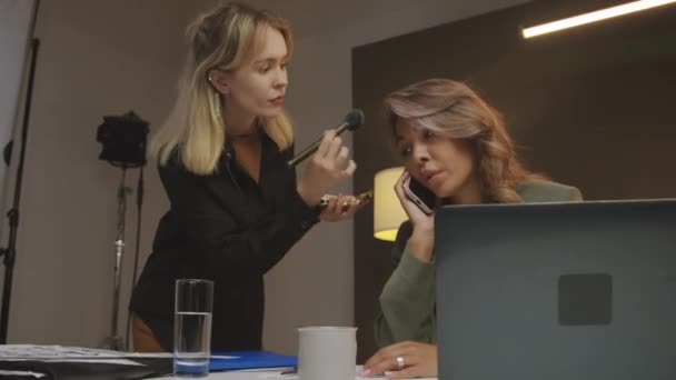 Μέση χαμηλή γωνία πλάνο του νεαρού καλλιτέχνη μακιγιάζ προσεκτικά powdering με μεγάλο πρόσωπο βούρτσα της γυναίκας παρουσιάστρια, ο οποίος κάθεται στο γραφείο, μιλώντας στο κινητό τηλέφωνο, ενώ ετοιμάζεται για συνέντευξη στο στούντιο - Πλάνα, βίντεο