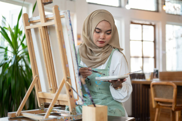 A talented Asian-Muslim female artist focuses on painting on canvas, mixing colors on a palette, working in her home studio. - Photo, Image