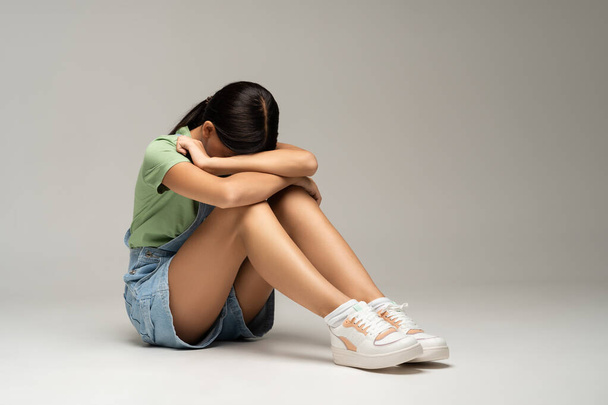 Frustrated feeling desperate teen girl closing face with hands sitting on gray background. Teenager living through loss of love, heartbreaking. Emotional pain, teenage crisis, depression concept. - Photo, Image