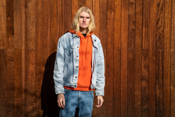 Apatyczny blond długie włosy człowiek skandynawski androgynous wygląd stoi w pobliżu drewnianego ogrodzenia patrząc w kamerę. LGBT, gej, transgenderowy biseksualista pozujący na naturę. Koncepcja osobowości indywidualnej. - Zdjęcie, obraz