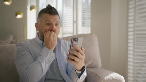Portrait of overjoyed comic man screaming while watching results of sport game on smartphone. Excited hilarious guy sitting on sofa in living room. Bearded male clapping and screaming with excitement - Footage, Video