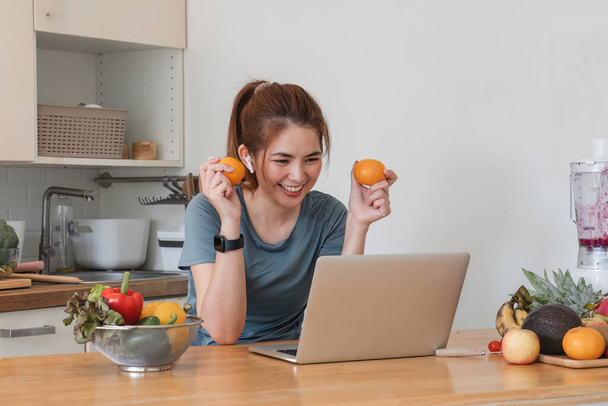 Gyönyörű fiatal nő gyakorló ruhák szórakozás egy aranyos konyha otthon. A laptop segítségével elkészíti a vegán gyümölcssaláta öntet, gyümölcs shake, vagy egészséges smoothies.. - Fotó, kép