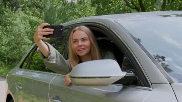 Exited smiling woman making video call with mobile phone from car window. Slow motion Local solo travel on weekends concept. Young traveler explore freedom outdoors in forest taking selfie photo - Footage, Video