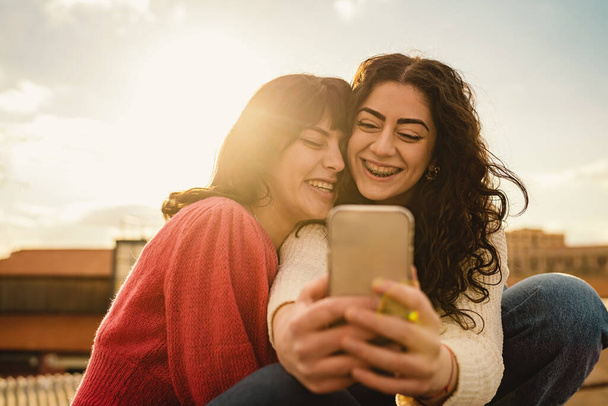 Due donne, radiosamente ridenti, condividono un momento con uno smartphone. Il sole tramonta dietro, gettando una tonalità dorata, incapsulando la loro gioia genuina e profonda connessione.. - Foto, immagini