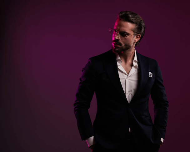 sexy elegant man in suit with open collar shirt holding hands in pockets and looking to side in front of purple background - Фото, изображение