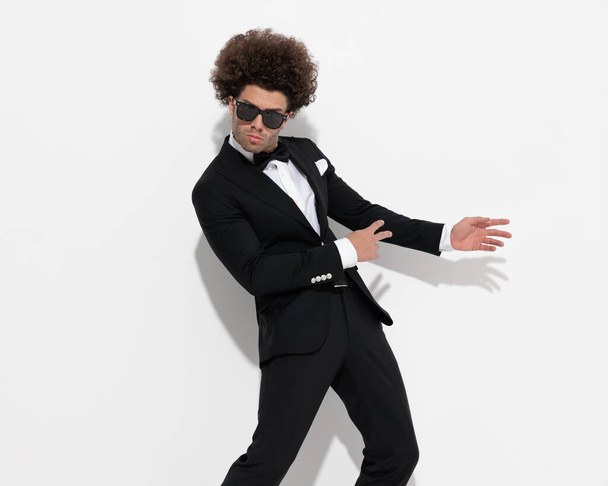 elegant young groom with curly hair in black tuxedo holding hands to side and showing in front of grey background - Photo, Image
