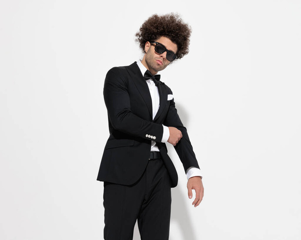 sexy curly hair guy with sunglasses adjusting tux jacket and being cool while posing in front of grey background - Фото, изображение
