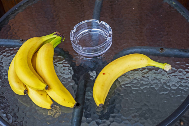 Een stel bananen en een banaan op een glazen ronde tafel in een café samen met een asbak. rookruimte - Foto, afbeelding