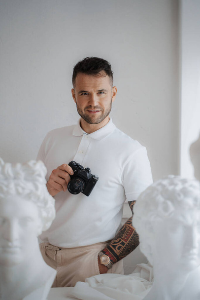 Hombre rico en una camisa blanca luciendo un tatuaje, posa con una cámara entre esculturas griegas antiguas en una habitación suavemente iluminada - Foto, Imagen