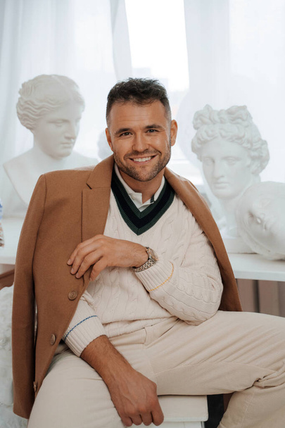 Portrait of a handsomely dressed man in old-money style, flashing a charming smile amid ancient Greek sculptures in a softly lit room - Photo, Image