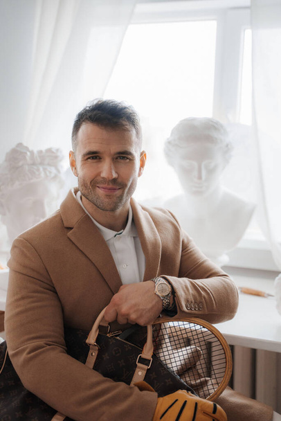 Charmanter Mann im alten Geld-Stil mit einem bezaubernden Lächeln, der eine Tasche und einen Tennisschläger inmitten antiker griechischer Skulpturen in einem Bildhaueratelier hält - Foto, Bild