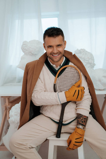 Charming man in old money style, sporting a captivating smile, poses with a tennis racket amid ancient Greek sculptures - Photo, Image