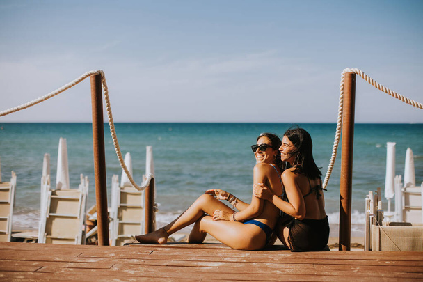 Δύο χαμογελαστές νεαρές γυναίκες με μπικίνι να κάθονται και να απολαμβάνουν διακοπές στην παραλία - Φωτογραφία, εικόνα