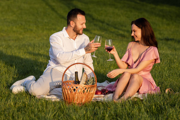 Shot of beautiful couple in park picnic outdoors. Loving married family drinking red wine sitting on blanket. Romantic date outside. Happy family enjoy drinks grapevine at nature. High quality photo - Photo, Image