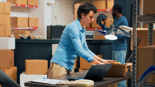 Small business owner putting supplies in cardboard boxes, shipping merchandise to customers. Young woman preparing shipment with warehouse goods, retail and logistics. Tripod shot. - 写真・画像