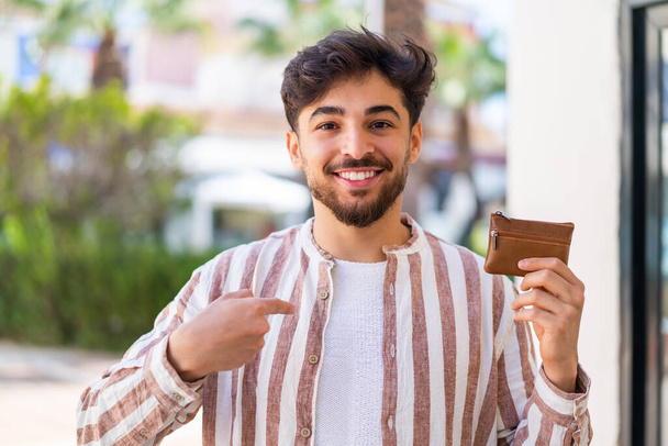 Hombre árabe guapo sosteniendo una billetera al aire libre con expresión facial sorpresa - Foto, Imagen