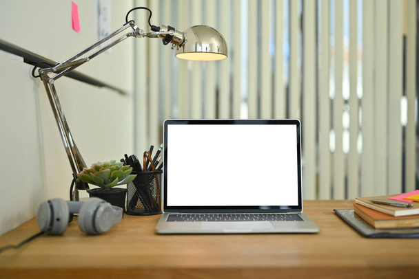Μπροστά όψη του φορητού υπολογιστή με λευκή οθόνη, ακουστικά, βιβλίο και λάμπα σε ξύλινο τραπέζι. - Φωτογραφία, εικόνα
