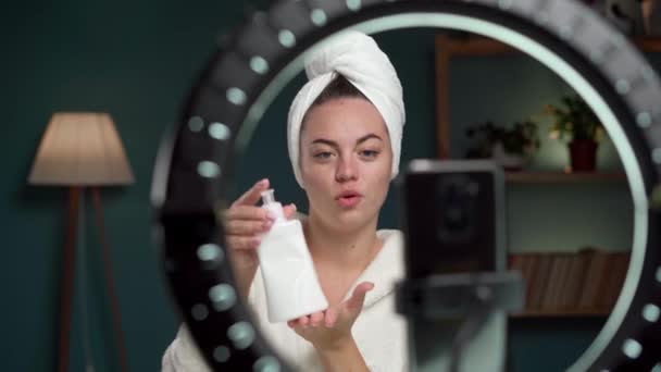 blogging, technology and people concept. Happy young blogger in bathrobe with ring light and smartphone streaming at home, talking about cosmetics product. Copy space - Footage, Video