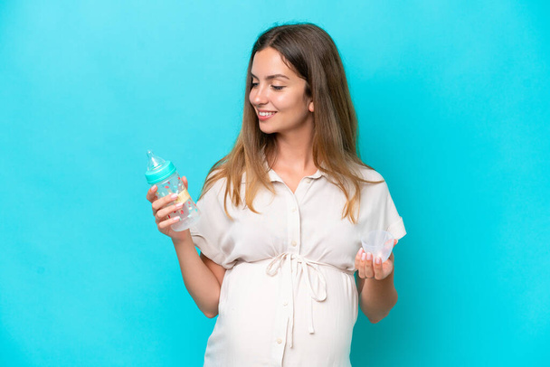 Молода біла жінка ізольована на синьому фоні вагітна і тримає пляшку для годування
 - Фото, зображення