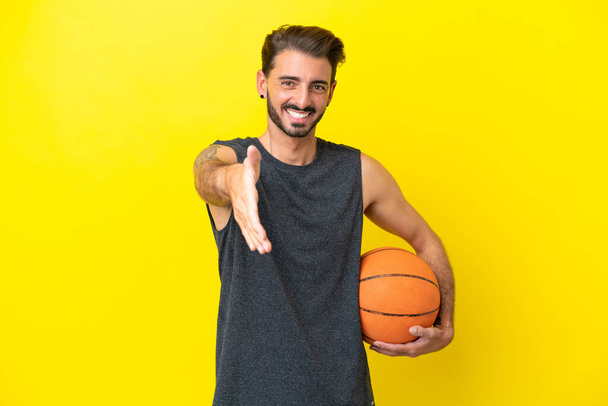 Красивый молодой баскетболист, изолированный на жёлтом фоне, пожимающий руку, чтобы заключить хорошую сделку - Фото, изображение