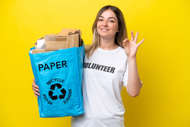 Junge rumänische Frau mit einer Recyclingtüte voller Papier zum Recyceln isoliert auf gelbem Hintergrund zeigt ok Zeichen mit den Fingern - Foto, Bild