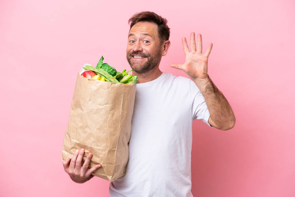 Mann mittleren Alters hält eine Einkaufstasche auf rosa Hintergrund isoliert und zählt mit den Fingern fünf - Foto, Bild