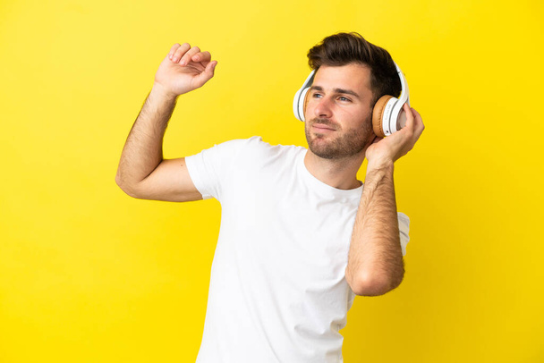 Νεαρός καυκάσιος όμορφος άνδρας απομονωμένος σε κίτρινο φόντο ακούγοντας μουσική και χορεύοντας - Φωτογραφία, εικόνα