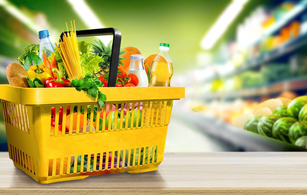 Food and groceries in shopping basket on wood table on blurred suppermarket aisle background - Photo, Image