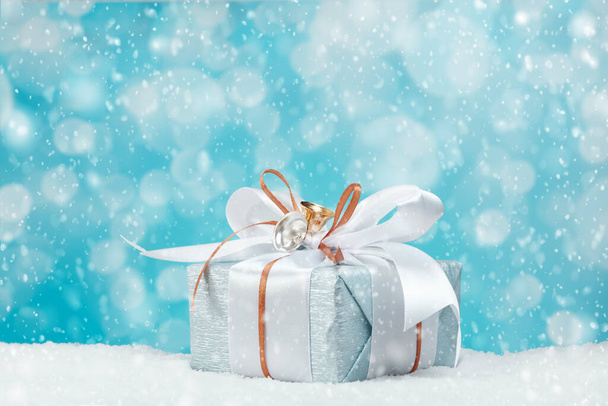 Gift box wrapped in blue silver paper with white and gold ribbon bow. Blue background, snow and bokeh light. Christmas and New Year gifts, Boxing Day. - Photo, Image