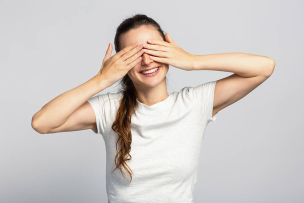 Χαρούμενη νεαρή ελκυστική γυναίκα που κρύβει τα μάτια της τα χέρια της, περιμένοντας την έκπληξη, στο ανοιχτό γκρι φόντο του στούντιο - Φωτογραφία, εικόνα