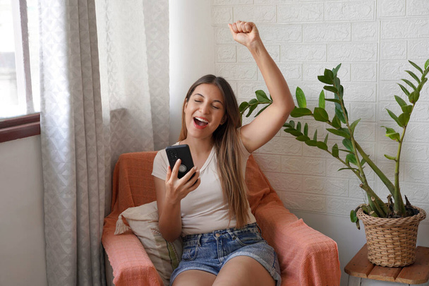 Jovem latina feliz segurando celular sentado em poltrona em casa. Feliz satisfeito feminino levanta o punho com smartphone na mão. - Foto, Imagem