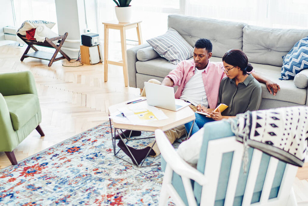 Young african american family resting together at free time in modern designed living room with stylish furniture, dark skinned couple in love watching online movie on laptop computer at home - Photo, Image