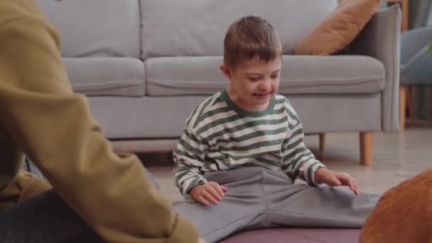 Χαρούμενος 6χρονος Καυκάσιος με σύνδρομο Down κάθεται στο χαλί στο άνετο σαλόνι παίζοντας με υπέροχο σκυλάκι και μαμά - Πλάνα, βίντεο