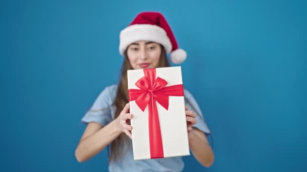 Young beautiful hispanic woman wearing christmas hat holding gift over isolated blue background - Footage, Video