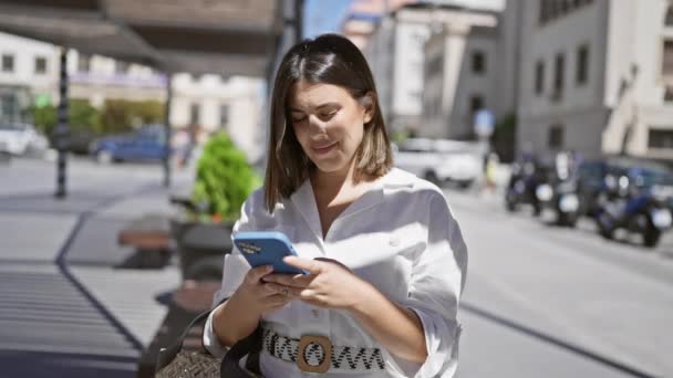 Νεαρή όμορφη ισπανόφωνη γυναίκα χαμογελά ευτυχισμένη χρησιμοποιώντας smartphone στην πόλη - Πλάνα, βίντεο