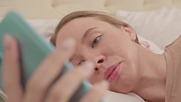 Közepes közelkép fiatal csinos kaukázusi nő böngészés okostelefonon, miközben feküdt nyugodtan az ágyban reggel - Felvétel, videó