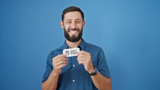 Jovem hispânico homem sorrindo confiante apontando para cartão de presente fazendo polegar acima gesto sobre fundo azul isolado - Filmagem, Vídeo