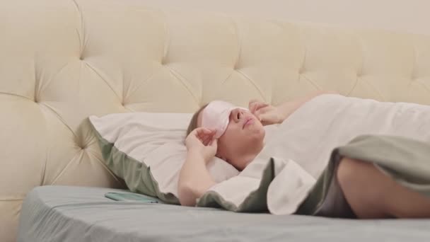 Mulher muito caucasiana decolando máscara de dormir e levando seu smartphone depois de acordar em cama confortável no hotel ou em casa de manhã - Filmagem, Vídeo